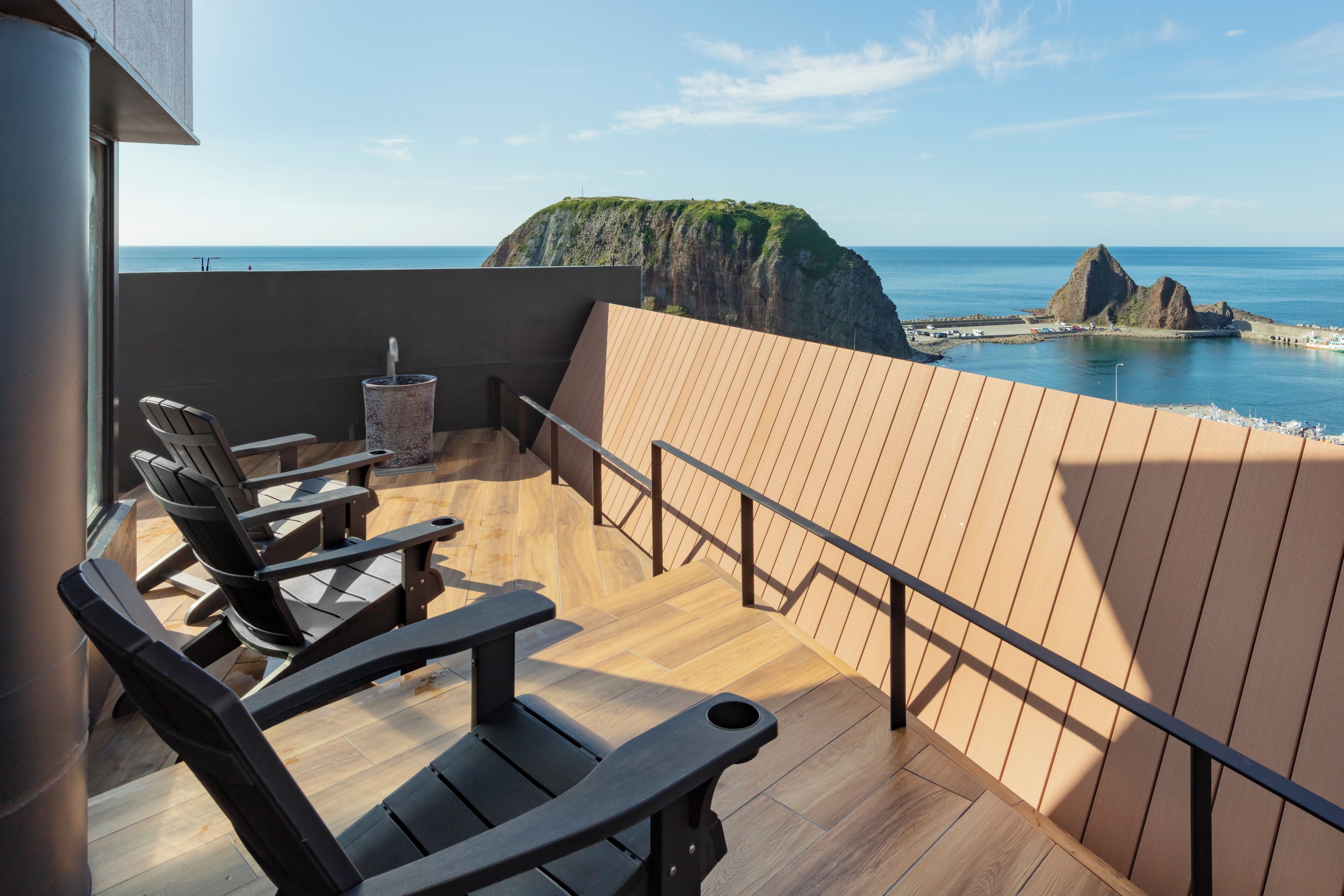 A wooden deck with chairs and a table, overlooking a coastal landscape with cliffs and the sea.
