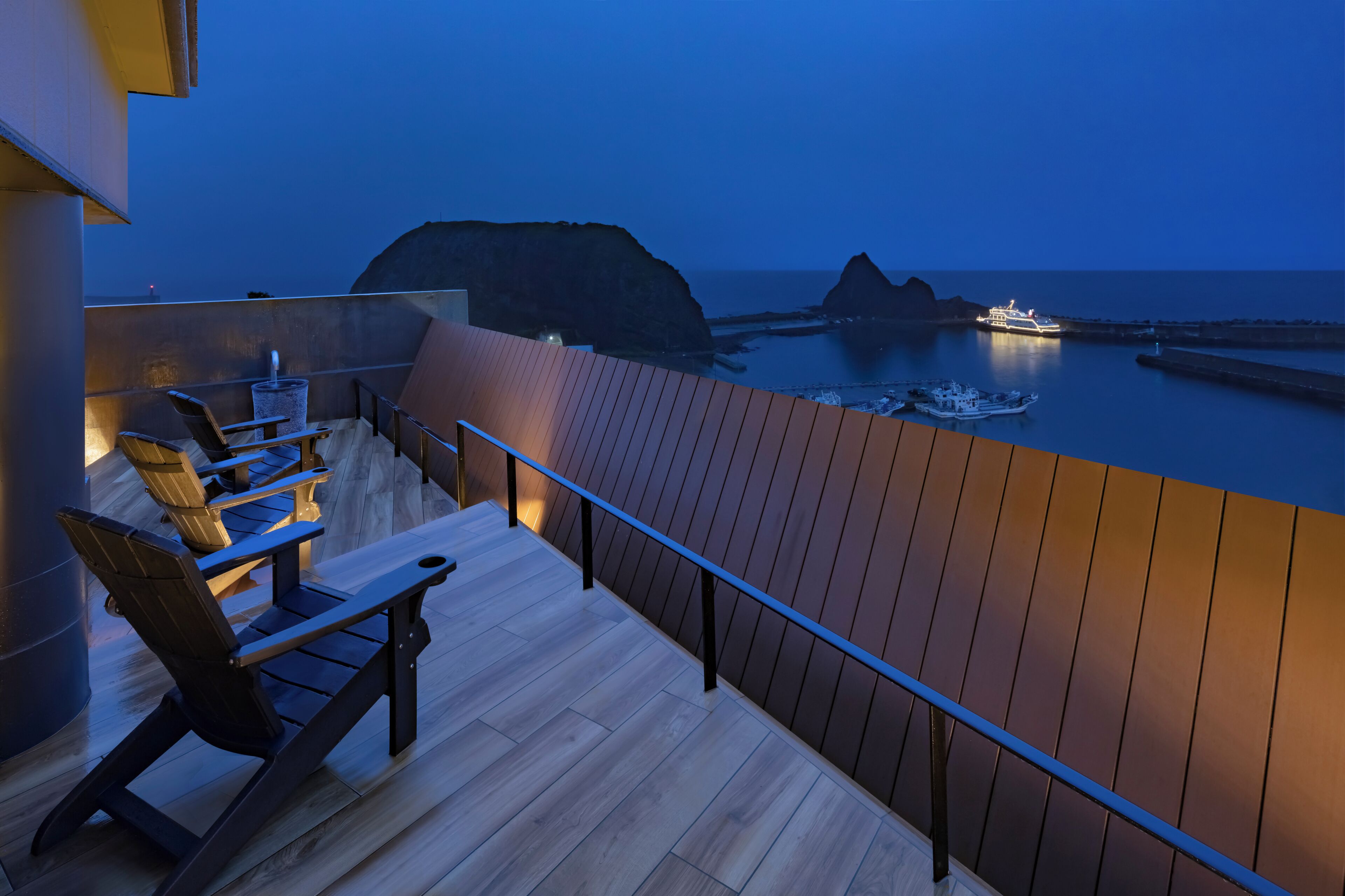 A balcony with wooden flooring and metal railings overlooking a calm sea and rocky coastline at dusk.