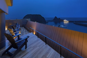 A balcony with wooden flooring and metal railings overlooking a calm sea and rocky coastline at dusk.