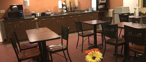 Uma área de refeições de hotel com mesas e cadeiras de madeira, uma máquina de café e uma TV no balcão.
