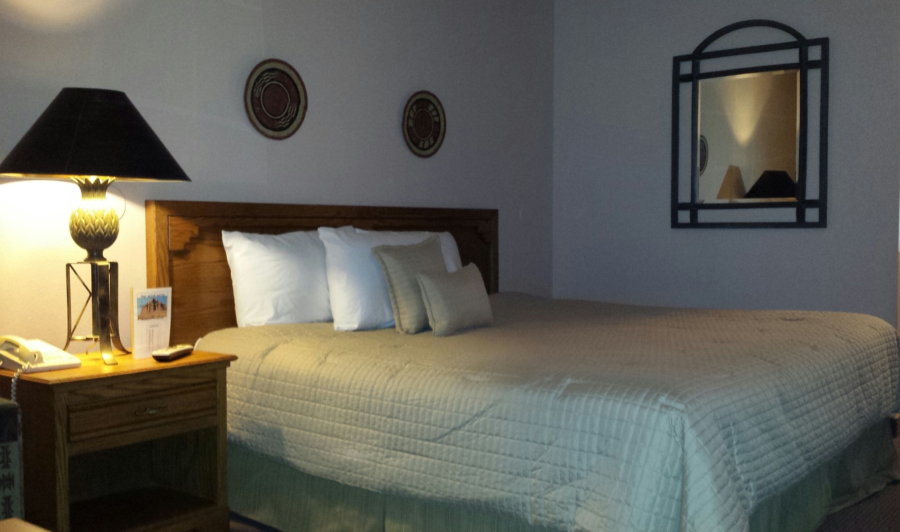 Una cama bien hecha con un edredón acolchado, una lámpara de noche, una mesita de noche con un teléfono y dos platos decorativos colgados en la pared.