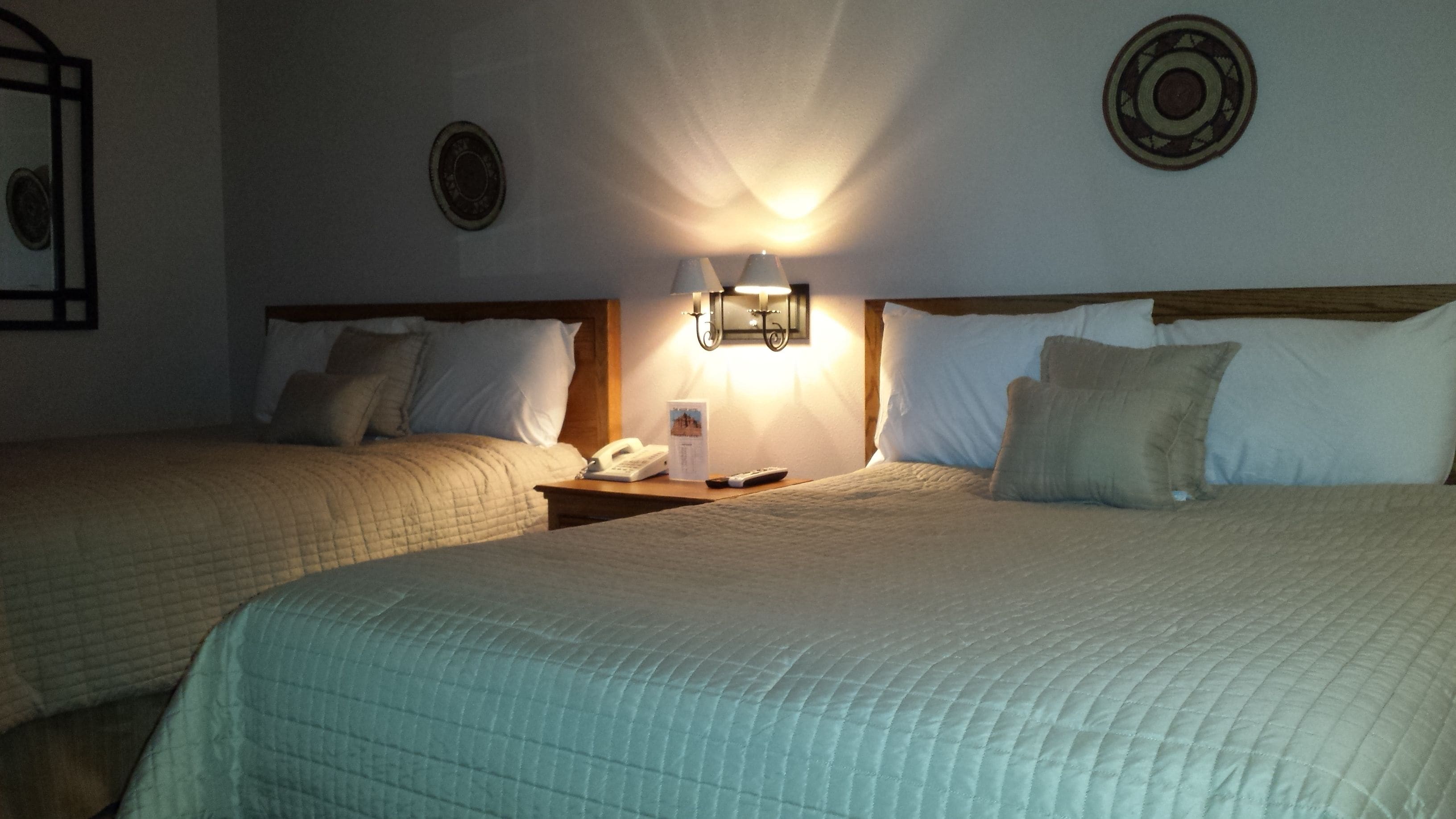 A hotel room with two beds, a bedside table with a phone and remote control, and a wall-mounted light fixture.