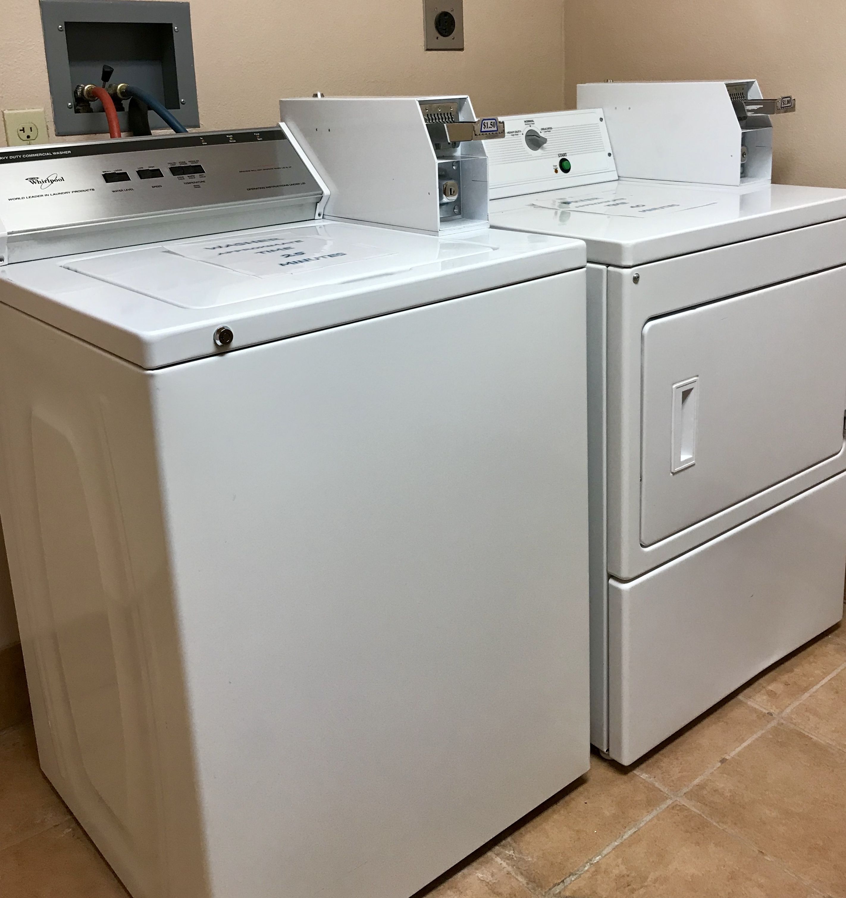 Ein Blick auf eine Waschküche mit zwei weißen Whirlpool Waschmaschinen und Trocknern, einer Steuerungstafel mit verschiedenen Einstellungen und einer Wasseranschlussdose über der Waschmaschine links.