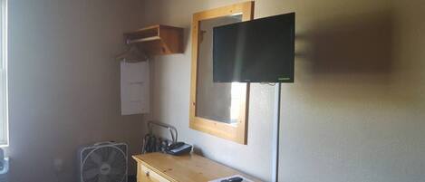 Ein kleines Zimmer mit einem Holzkasten, einem Schreibtisch, einem Spiegel, einem Fernseher und einem Ventilator.