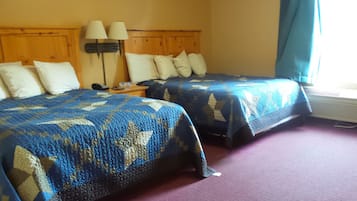 Two single beds with blue and beige patterned blankets, a wooden headboard, a nightstand with a lamp, and a window with a blue curtain.