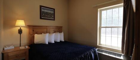 A hotel room with a bed, a nightstand, a lamp, a window with curtains, and a picture on the wall.