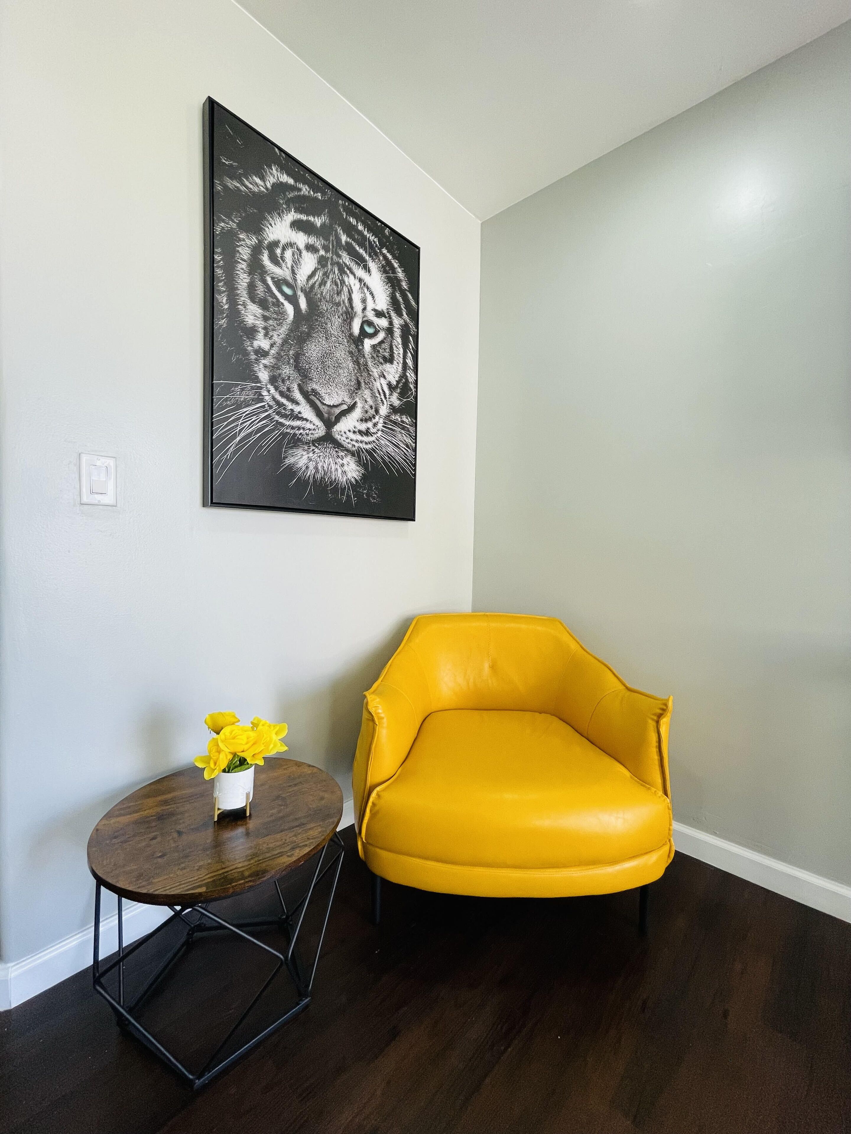 A room with a yellow armchair, a round wooden table with a vase of yellow flowers, and a black and white tiger painting on the wall.