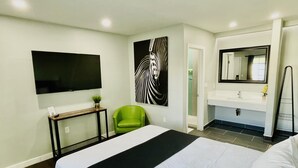 A hotel room with a large bed, a green chair, a flat-screen TV, a painting, a bathroom with a mirror, and a towel rack.