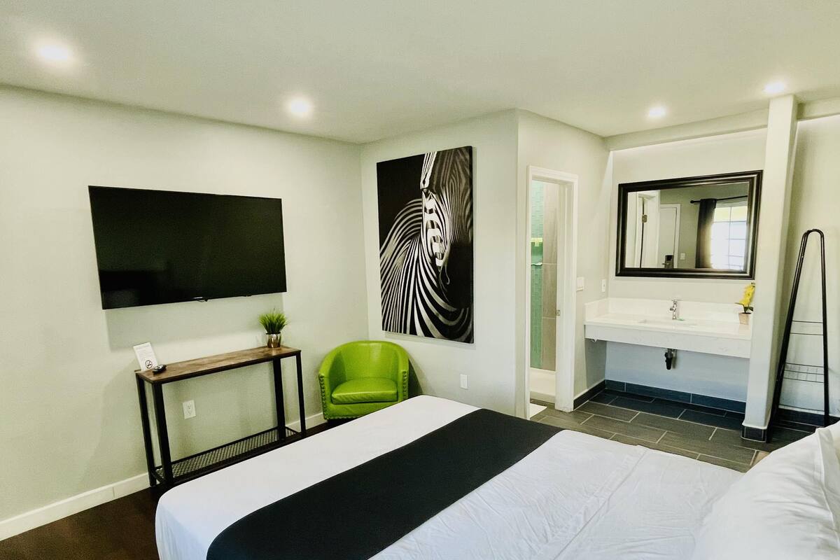A hotel room with a large bed, a green chair, a flat-screen TV, a painting, a bathroom with a mirror, and a towel rack.