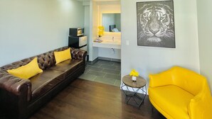 A modern living room with a brown leather sofa, a yellow armchair, and a small round table with a vase of yellow flowers. There is a kitchen area in the background with a microwave and a sink.