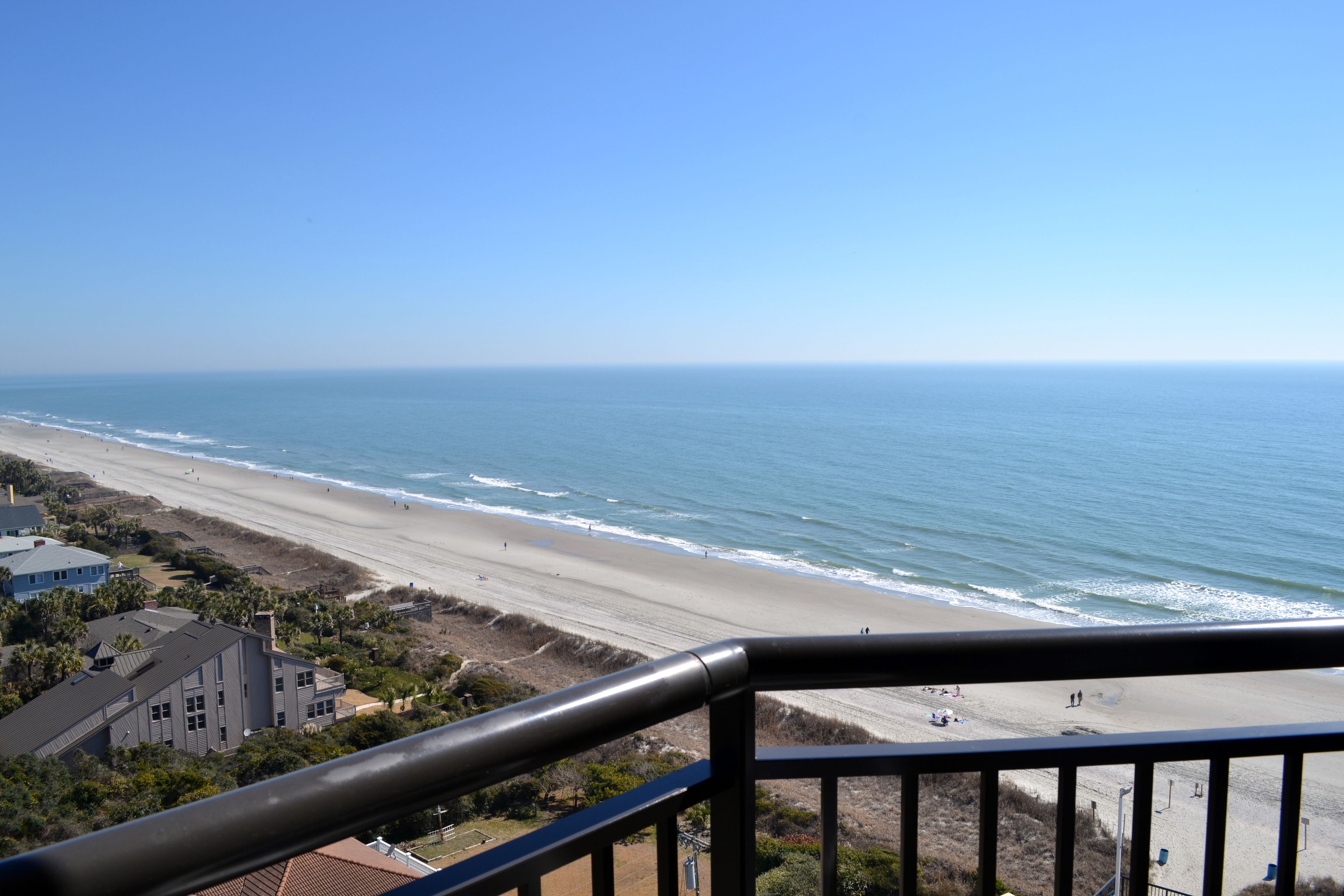 A beach view from a balcony with a railing, ocean, and buildings in the background.