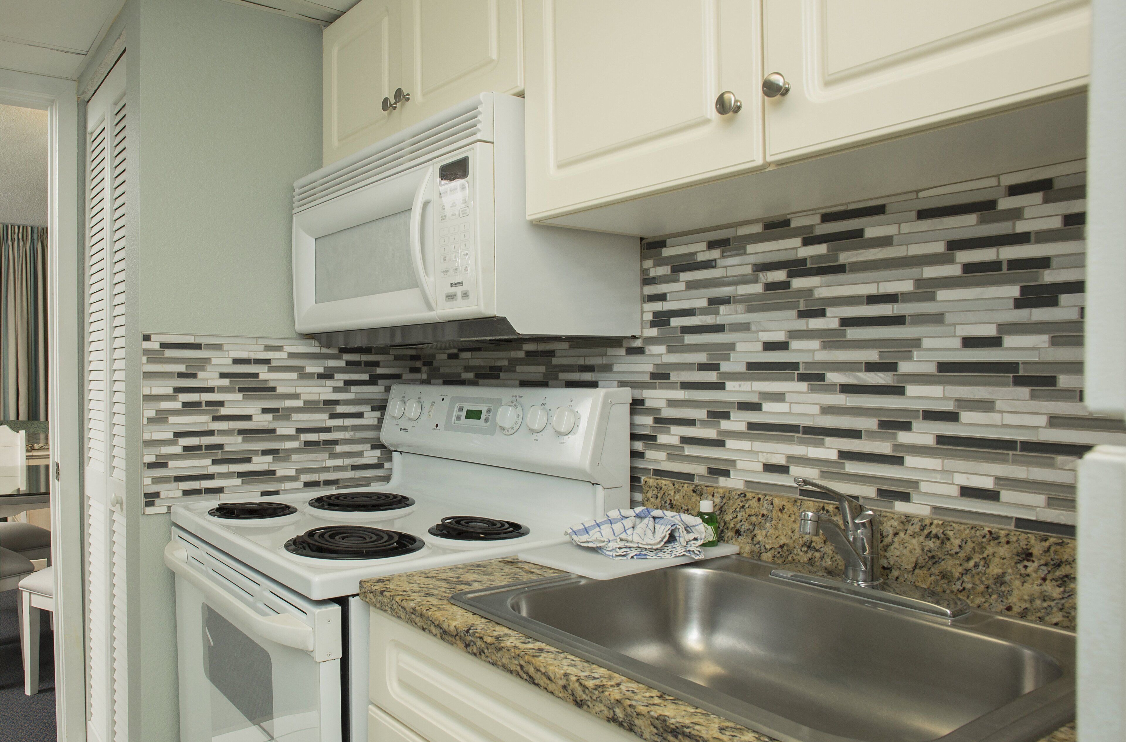 A kitchen with white cabinets, a microwave, stove, granite countertop, stainless steel sink, and a tiled backsplash.