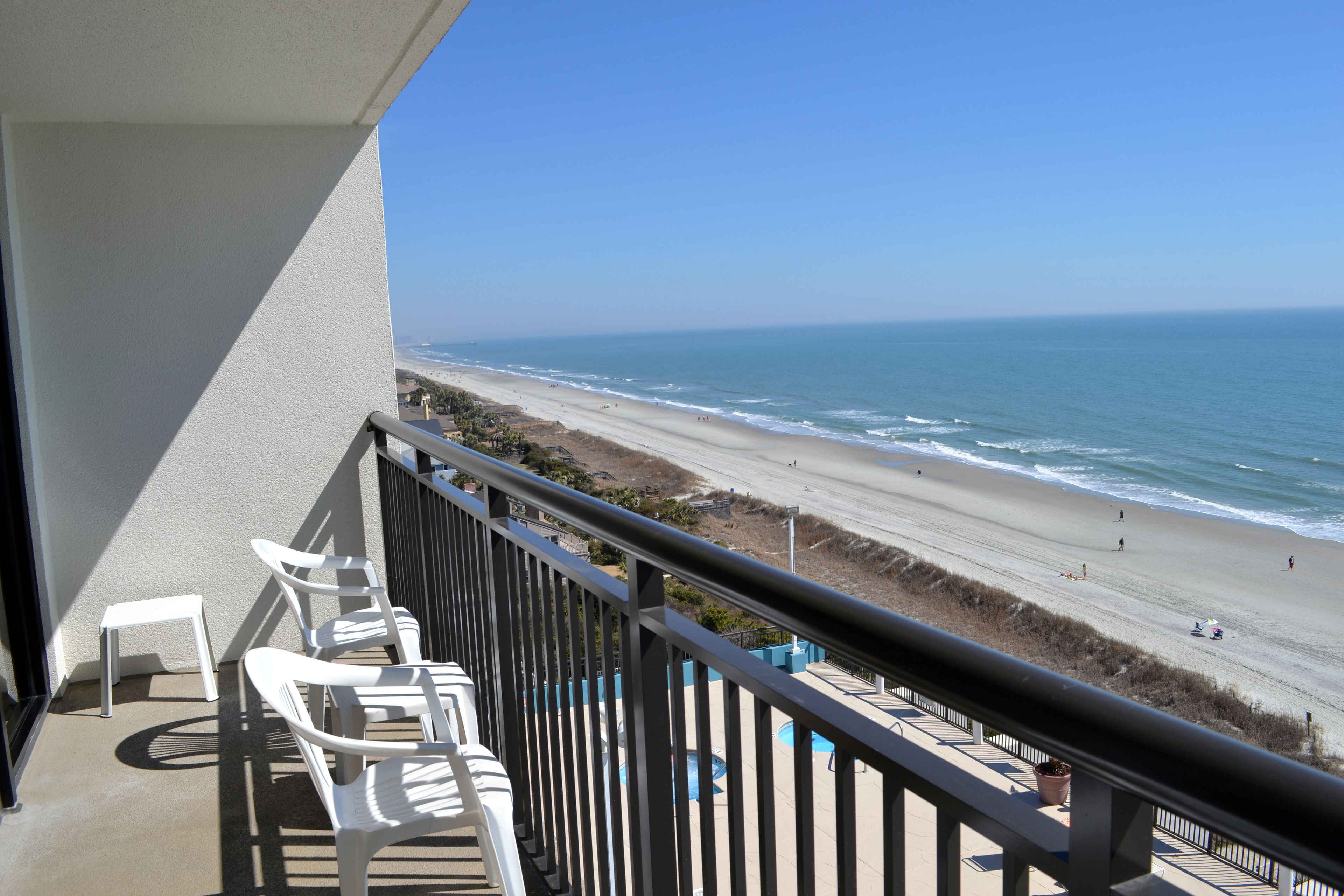 A balcony with white plastic chairs and a view of the beach and ocean.