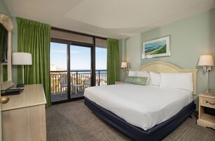 Habitación de hotel con una cama grande, dos mesitas de noche, un escritorio con lámpara, una silla y vista al océano a través de una puerta corrediza de vidrio.