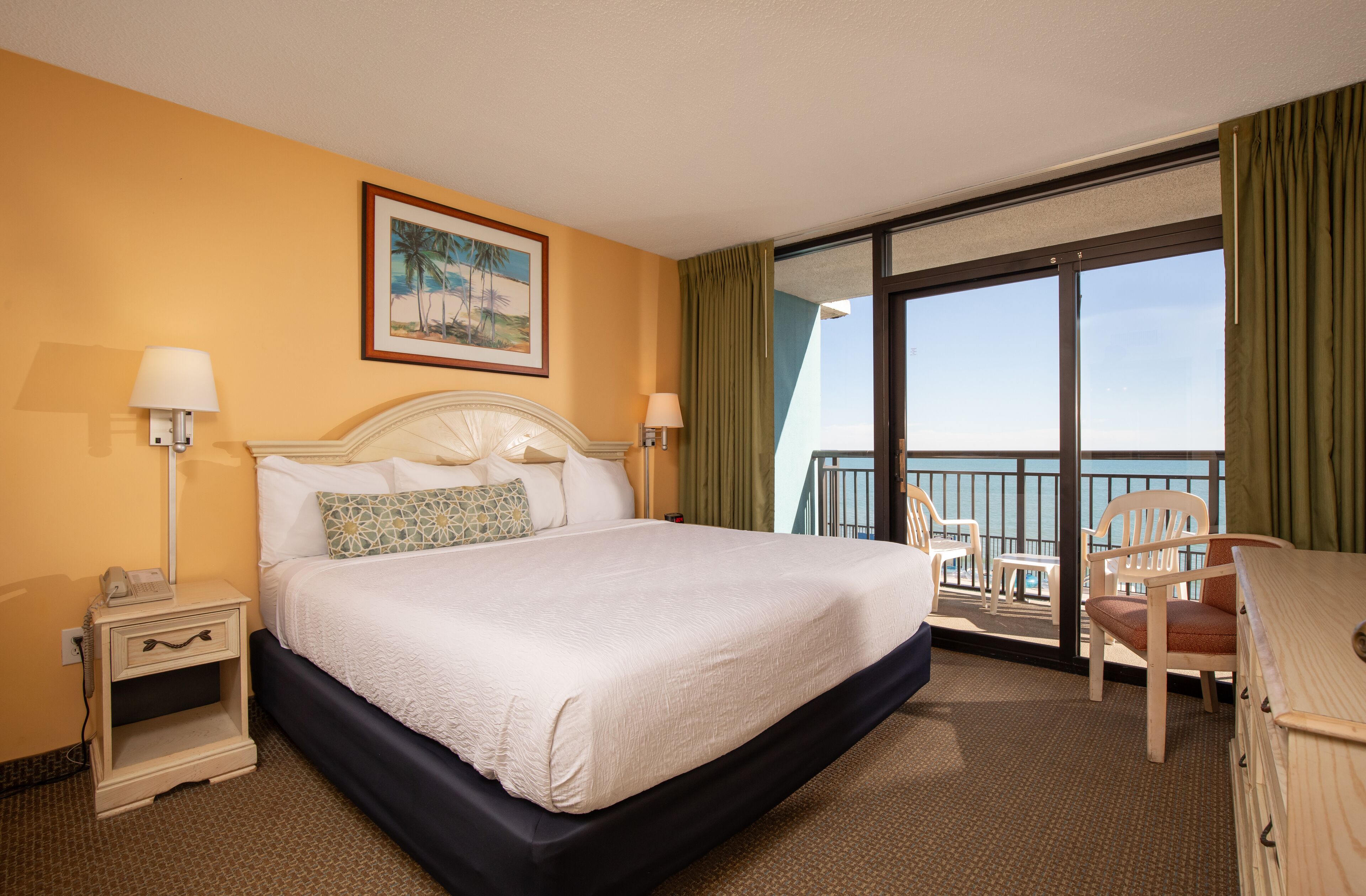A hotel room with a large bed, two bedside tables, a desk with a chair, a balcony with a view, and a painting on the wall.