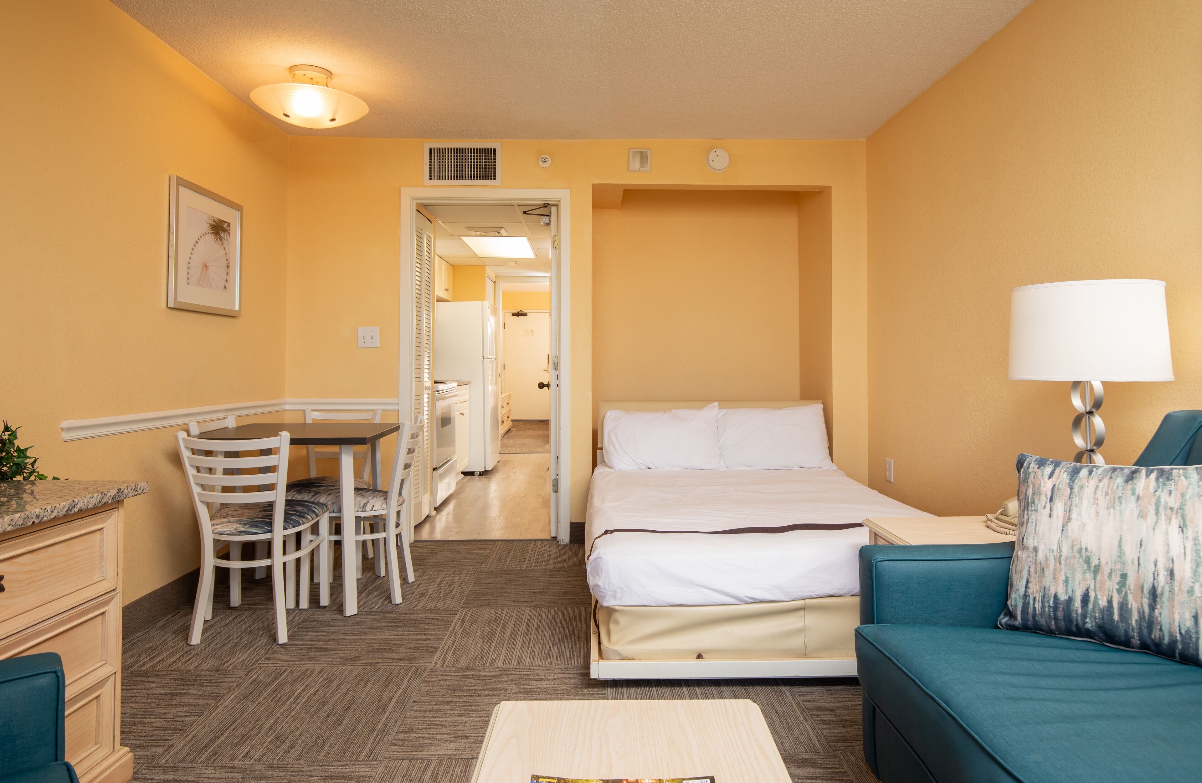 Habitación de hotel con cama, escritorio con sillas, lámpara y vista al pasillo.