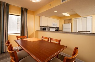 Un comedor con mesa y sillas de madera, una cocina con armarios y electrodomésticos blancos, y un ventanal con cortinas.