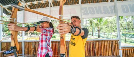 A person holding a bow and arrow, another person assisting with the bow, and a bamboo structure in the background.
