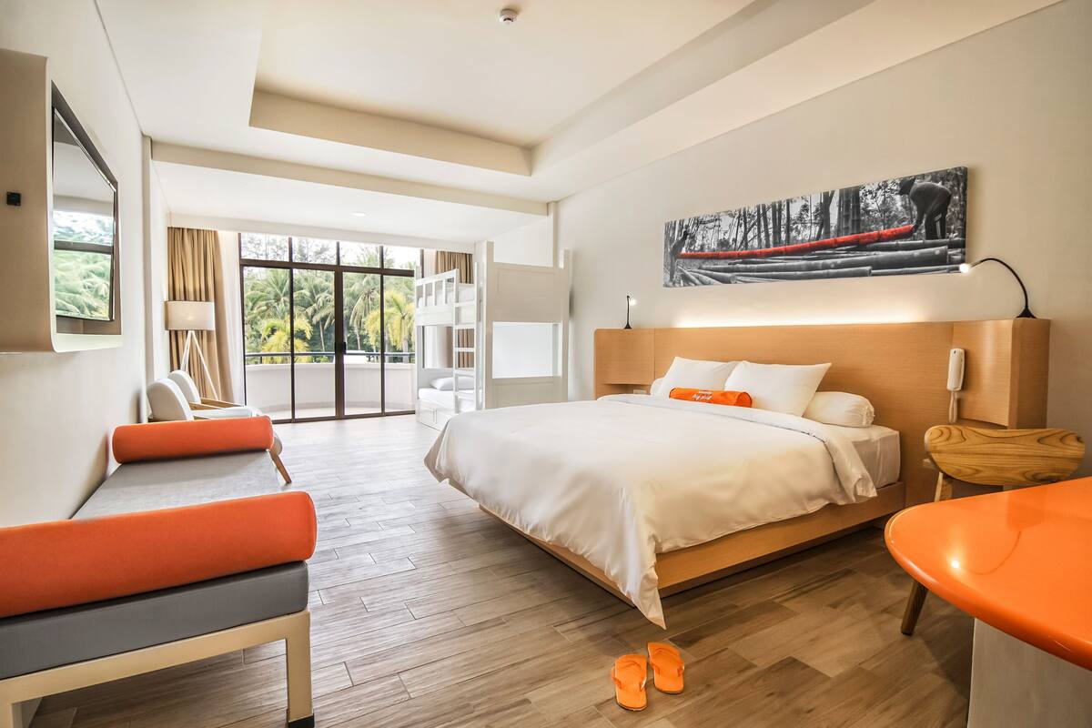 A modern hotel room with a large bed, a seating area with a bench and chairs, a small table, and a balcony with a view of palm trees.