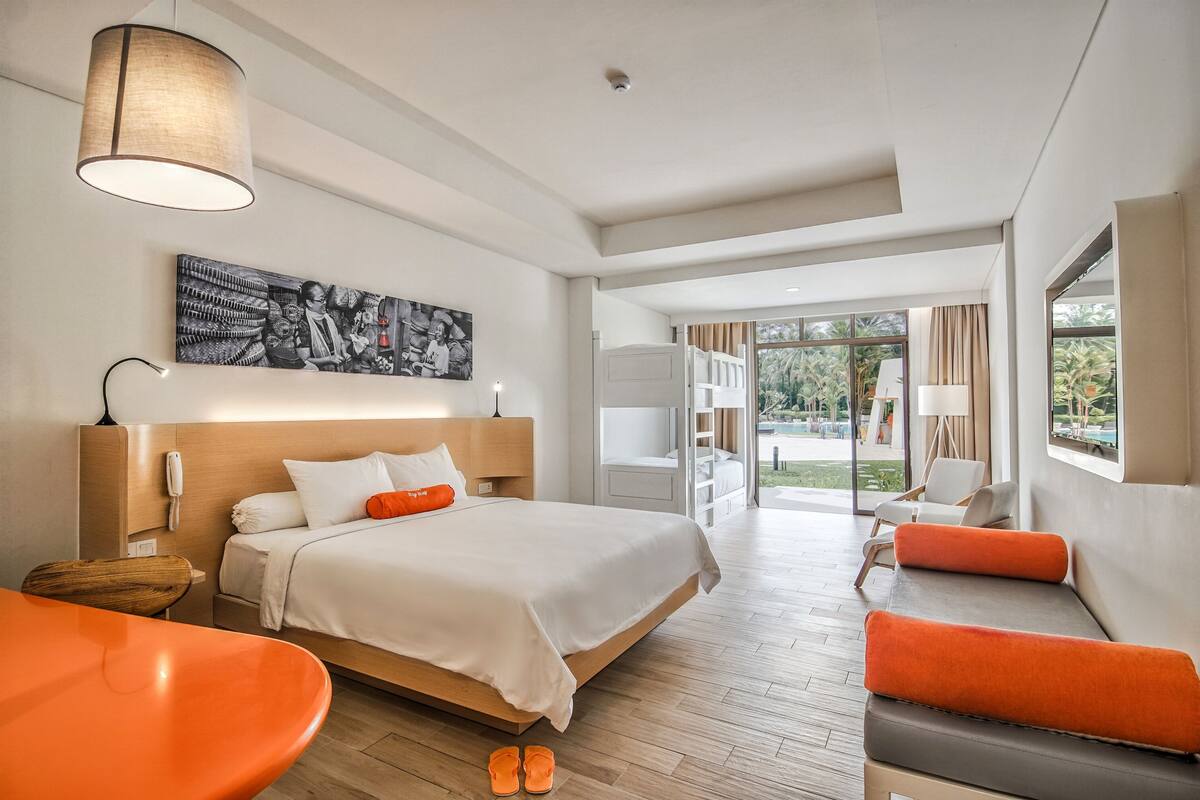 A modern hotel room with a large bed, a sitting area with orange furniture, and a view of the outdoors.