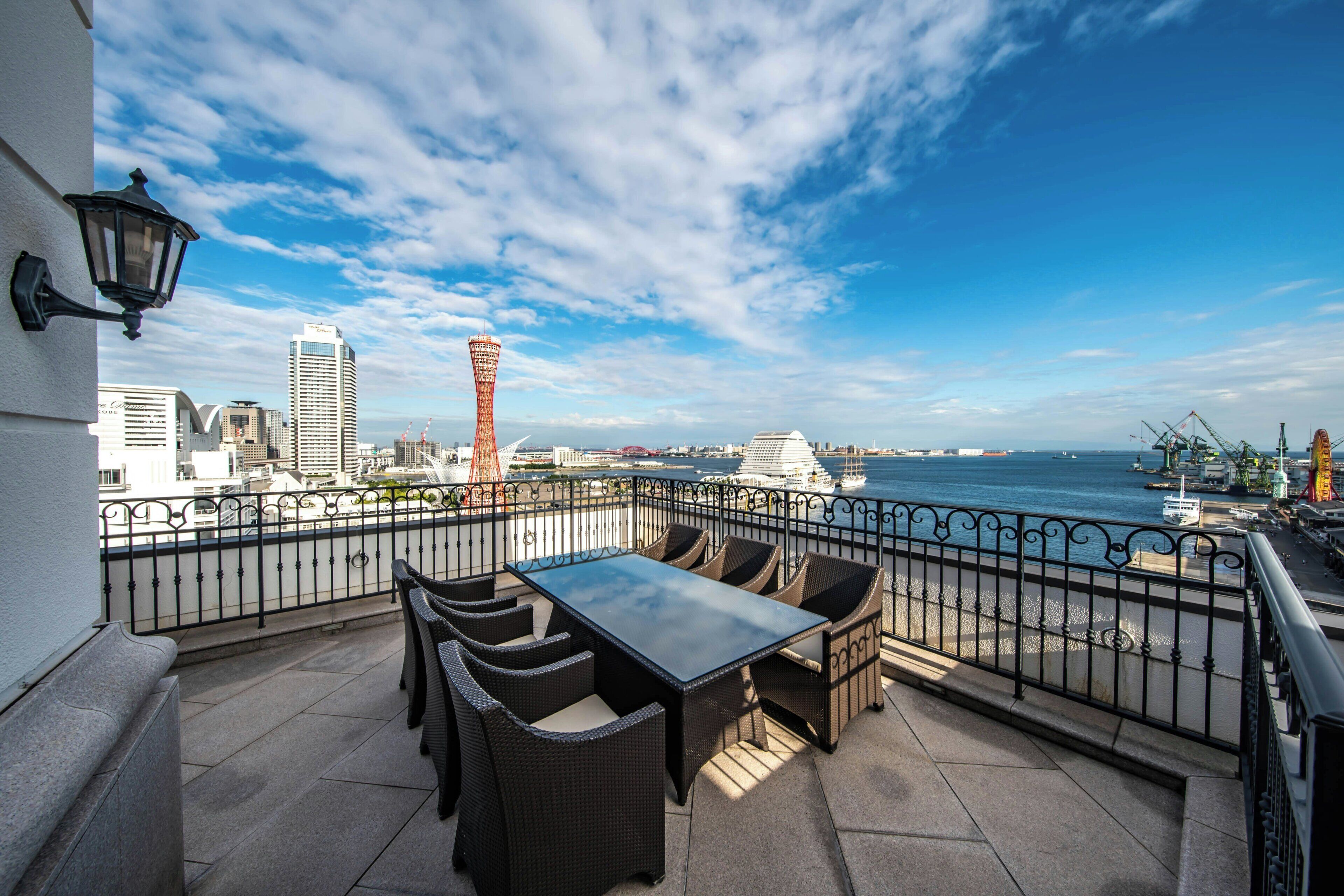 Un balcón con mobiliario exterior, vista al mar y edificios de la ciudad al fondo.