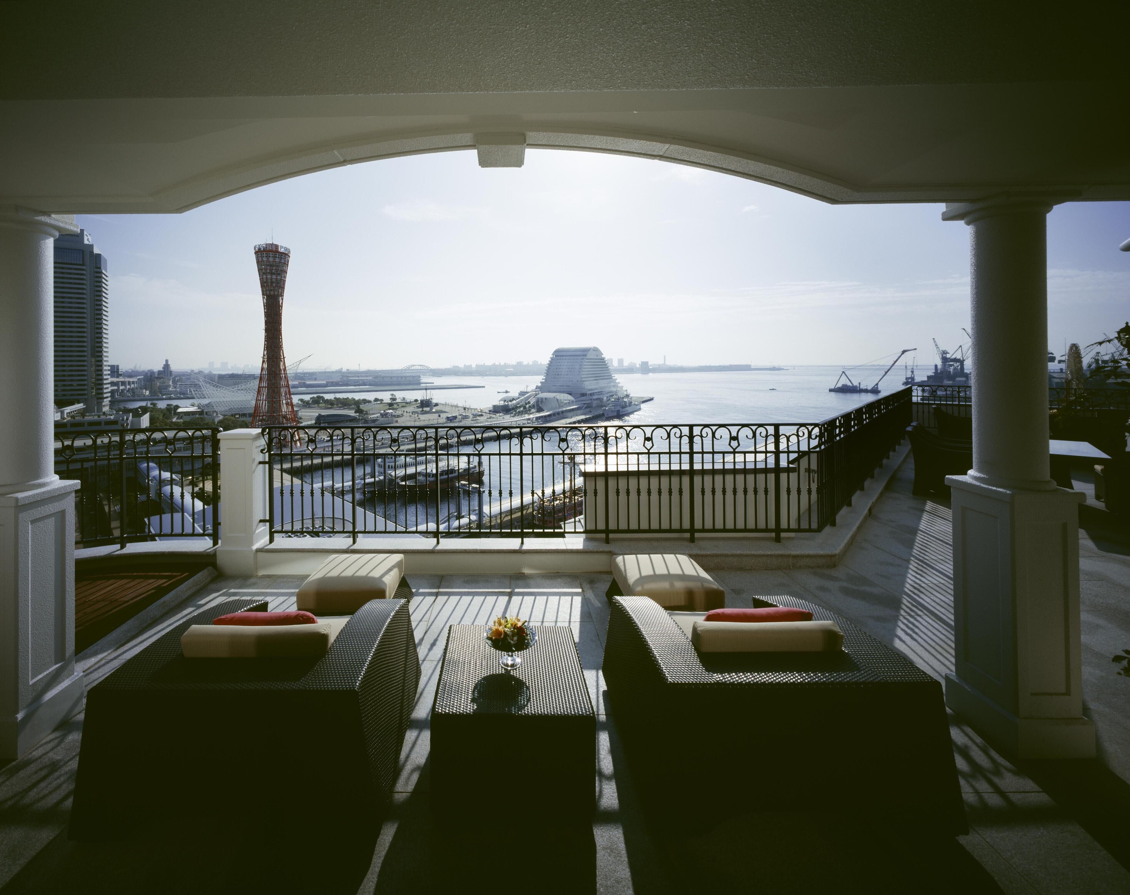 A balcony with outdoor seating, overlooking a waterfront with a tall tower and a cruise ship.