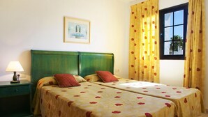 A room with two beds, a nightstand, a lamp, a framed picture on the wall, and a window with yellow curtains. - Apartamentos Flamingo (Tías)