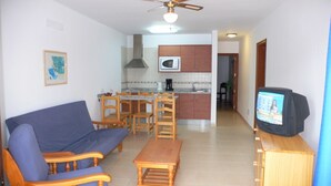A living room with a sofa, wooden chairs, a coffee table, a television, and a kitchen area with a microwave. - Apartamentos Flamingo (Tías)