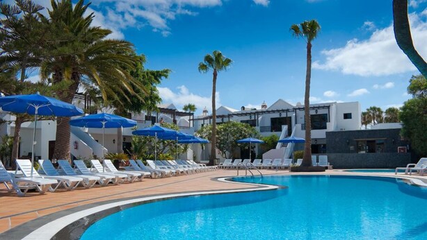 A swimming pool with lounge chairs and umbrellas, surrounded by palm trees and modern buildings. - Apartamentos Flamingo (Tías)