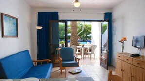 A room with a blue sofa, a dining table with plates and cups, a TV, and a view of an outdoor pool area. - Apartamentos Flamingo (Tías)