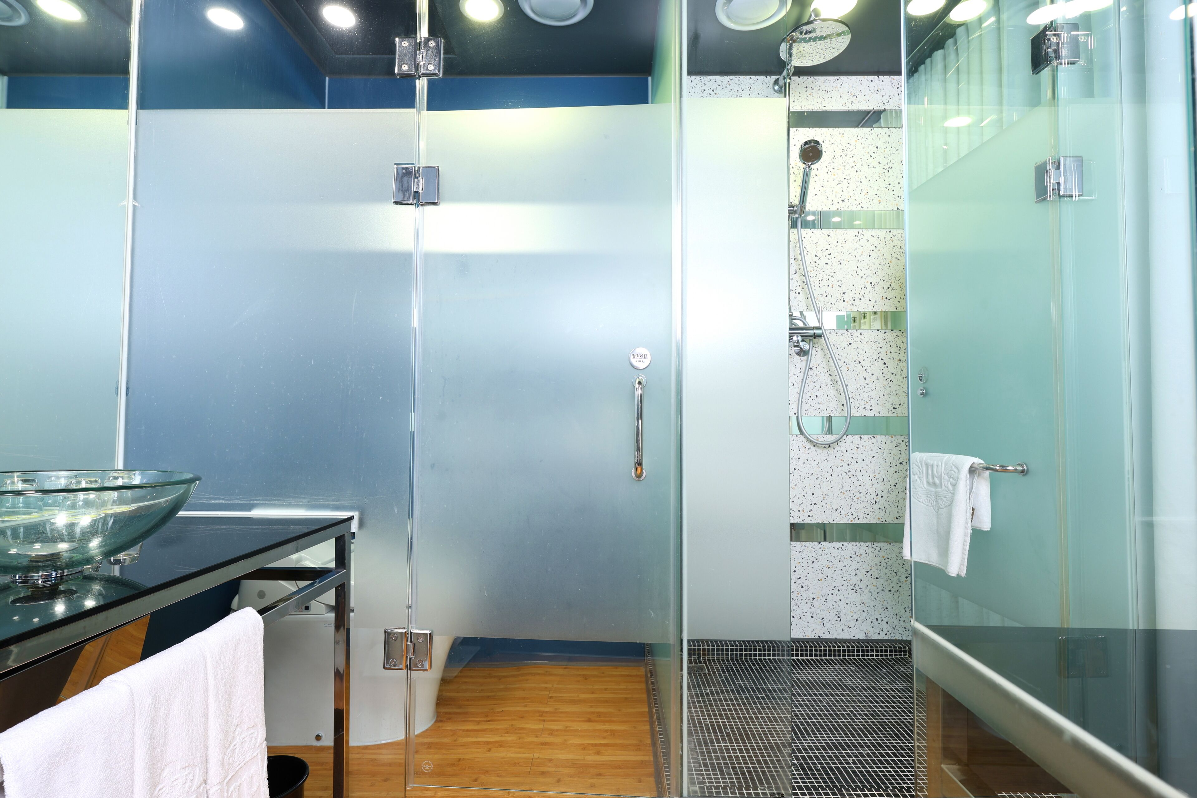 A modern bathroom with a glass shower enclosure, a glass sink, and a wooden floor.