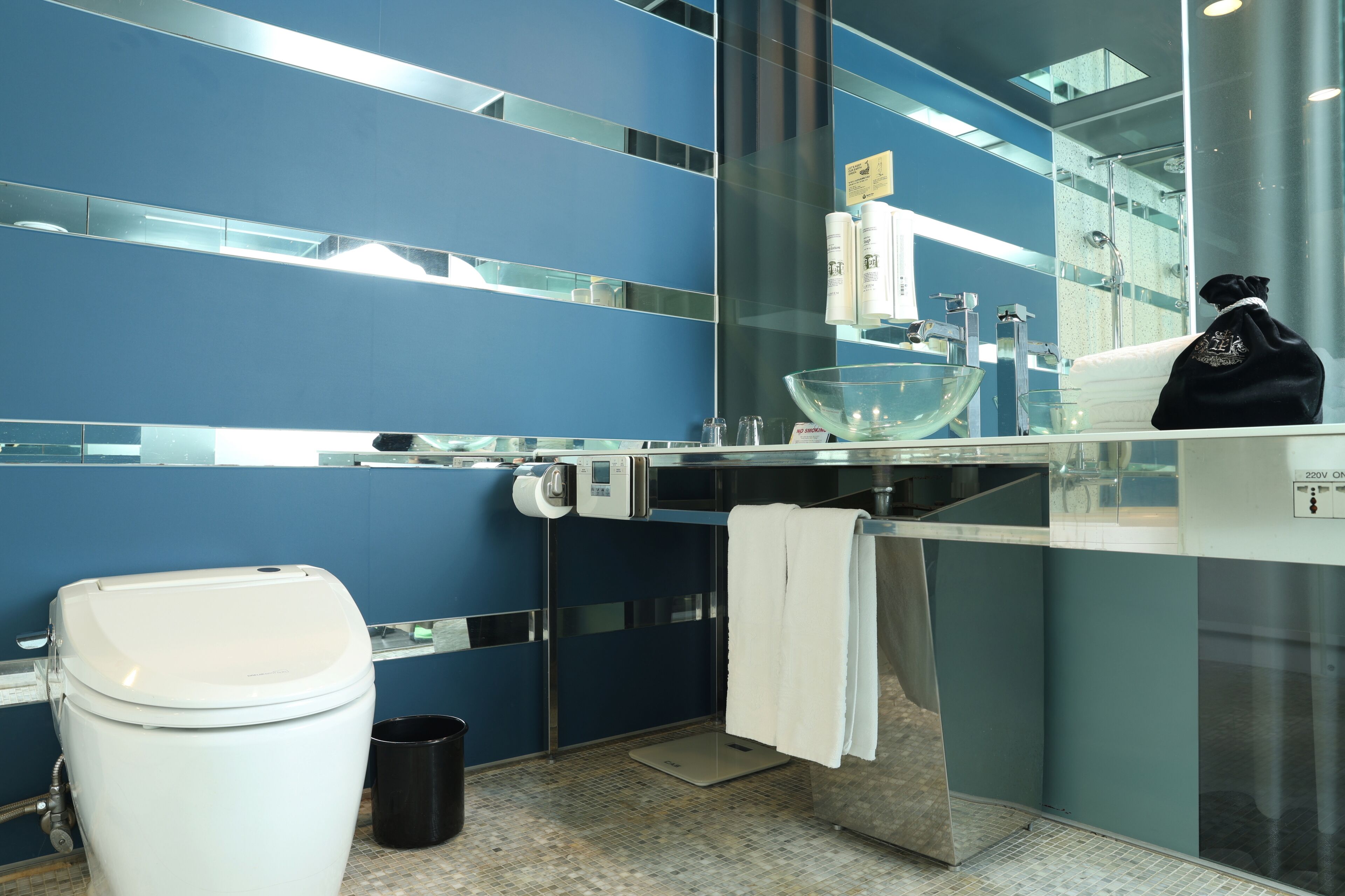 A modern bathroom with a blue tiled wall, a glass sink, a white toilet, and a black bag on the counter.