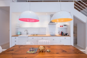 Une cuisine moderne avec des meubles blancs, une table à manger en bois et des suspensions lumineuses aux abat-jour rouges et oranges.