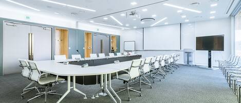 A conference room with a long table, white chairs, a whiteboard, and a large screen.