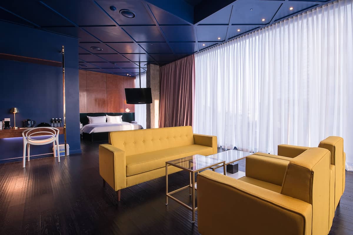 Une chambre d’hôtel moderne avec un canapé jaune, une table basse en verre, un lit avec du linge de maison blanc et une grande fenêtre ornée de rideaux légers.