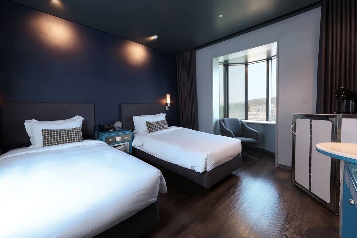Une chambre d’hôtel avec deux lits, un bureau, une chaise et une grande fenêtre donnant sur la ville.