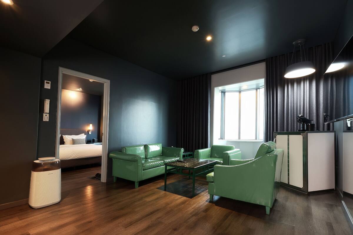 Une chambre d’hôtel moderne avec un lit, des canapés verts, une table basse et un bureau avec une lampe.