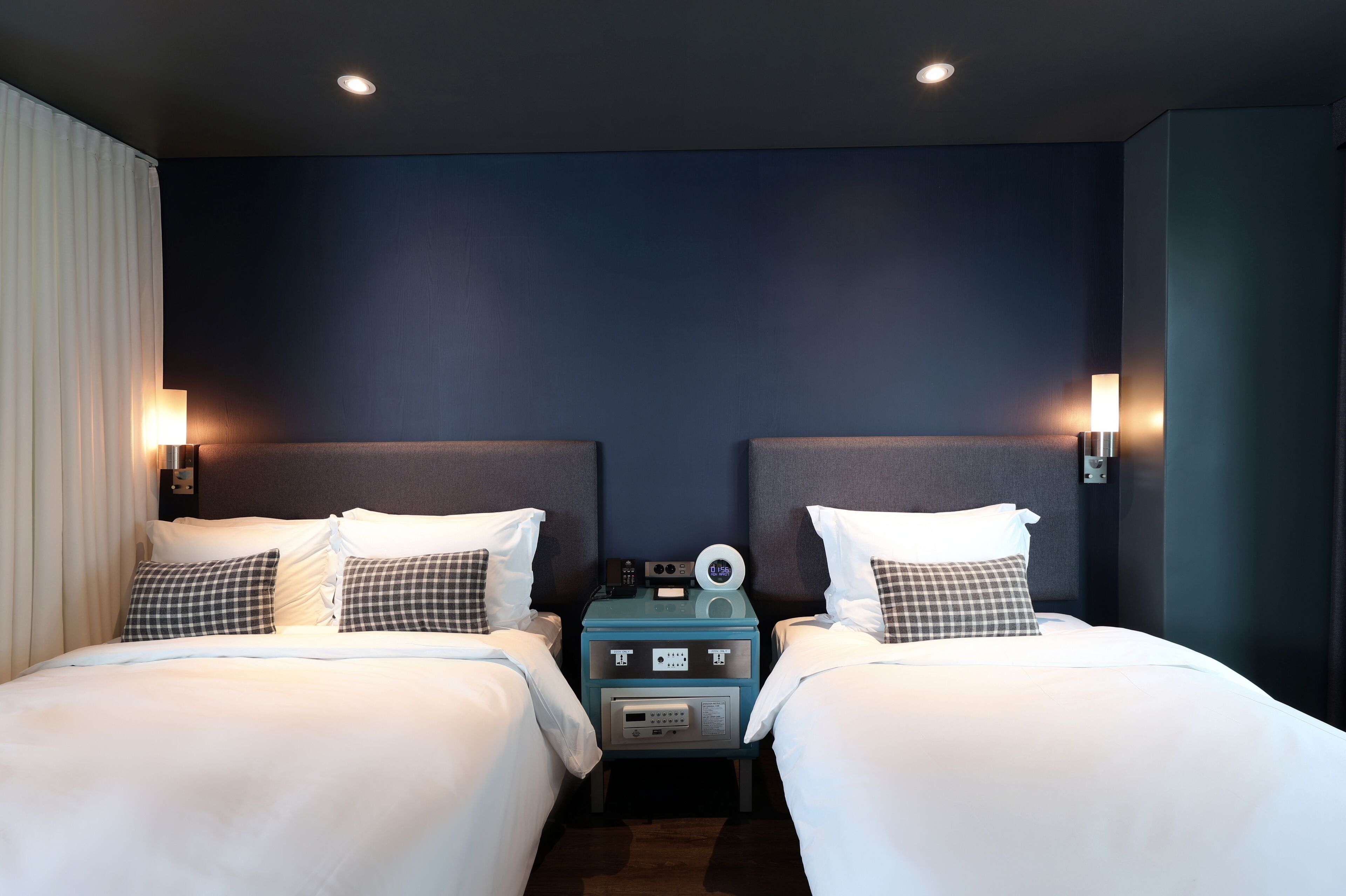 A hotel room with two beds, a nightstand with a clock and a lamp, and a dark blue accent wall.