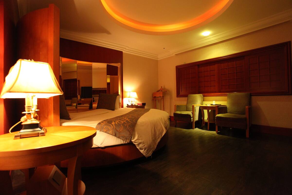 A hotel room with a bed, a desk with a lamp, two chairs, and a small table.