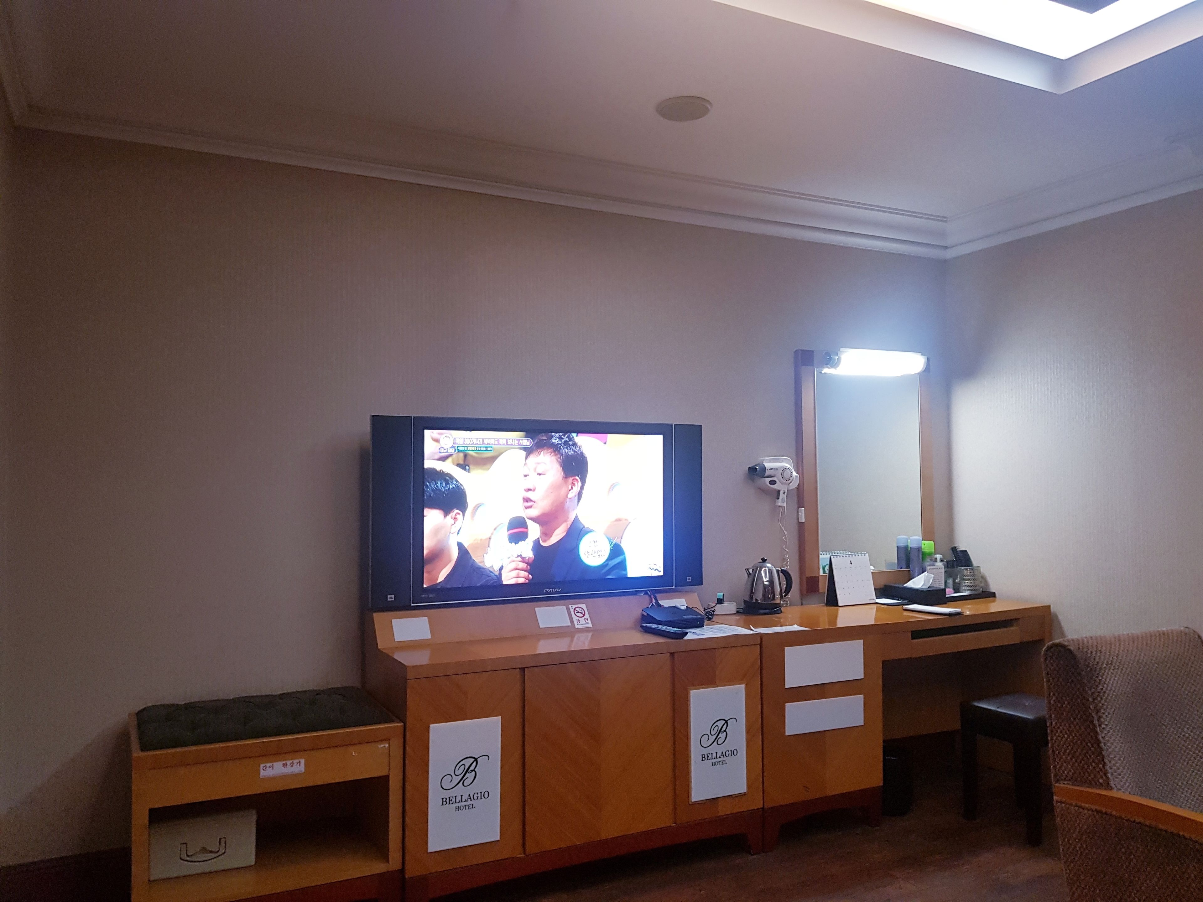 A hotel room with a television mounted on the wall, a desk with a chair, a mirror, and a small shelf with items.