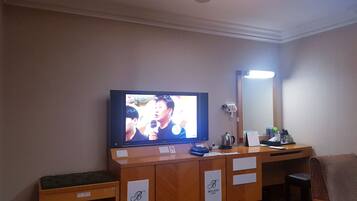 A hotel room with a television mounted on the wall, a desk with a chair, a mirror, and a small shelf with items.