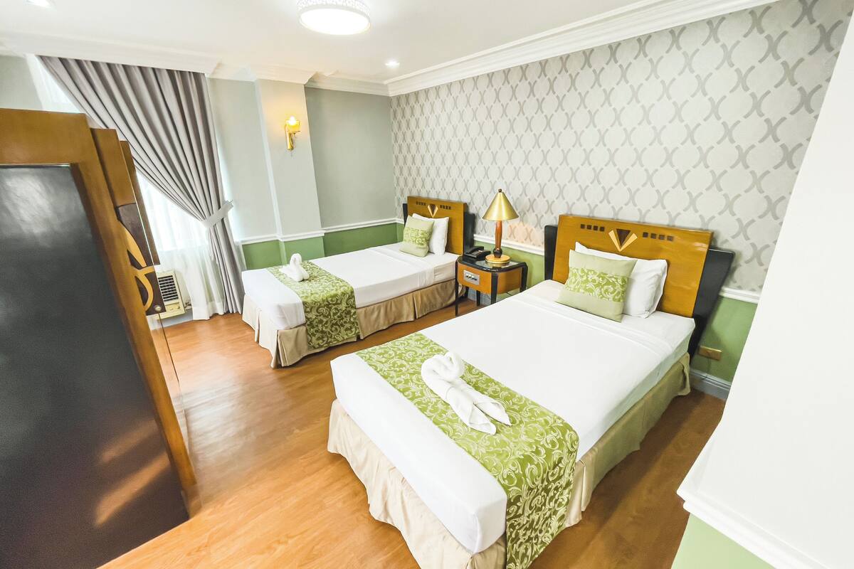 A hotel room with two beds, a nightstand, a lamp, and a window with curtains.