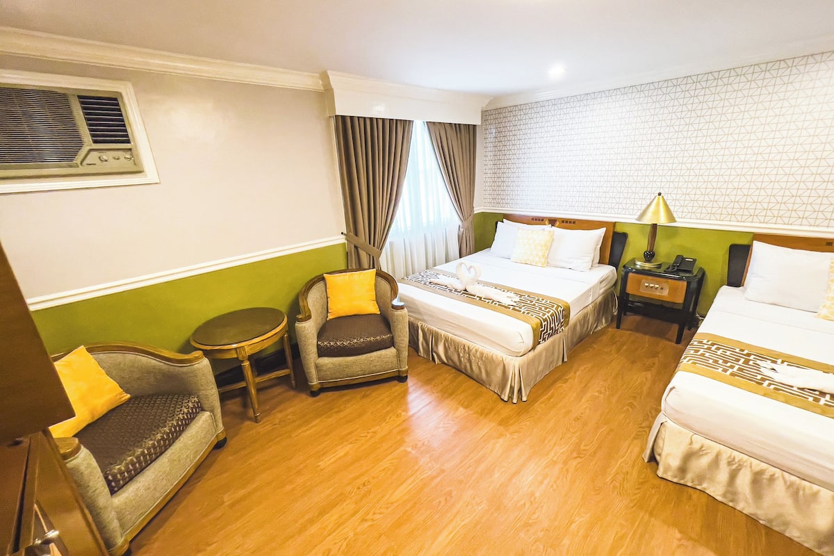 A hotel room with a bed, two armchairs, a small table, a nightstand, and a wall-mounted air conditioning unit.