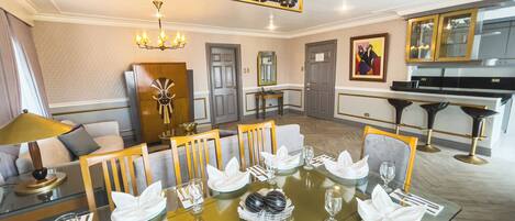 A dining area with a glass table set for a meal, a chandelier above, and a piano in the background.