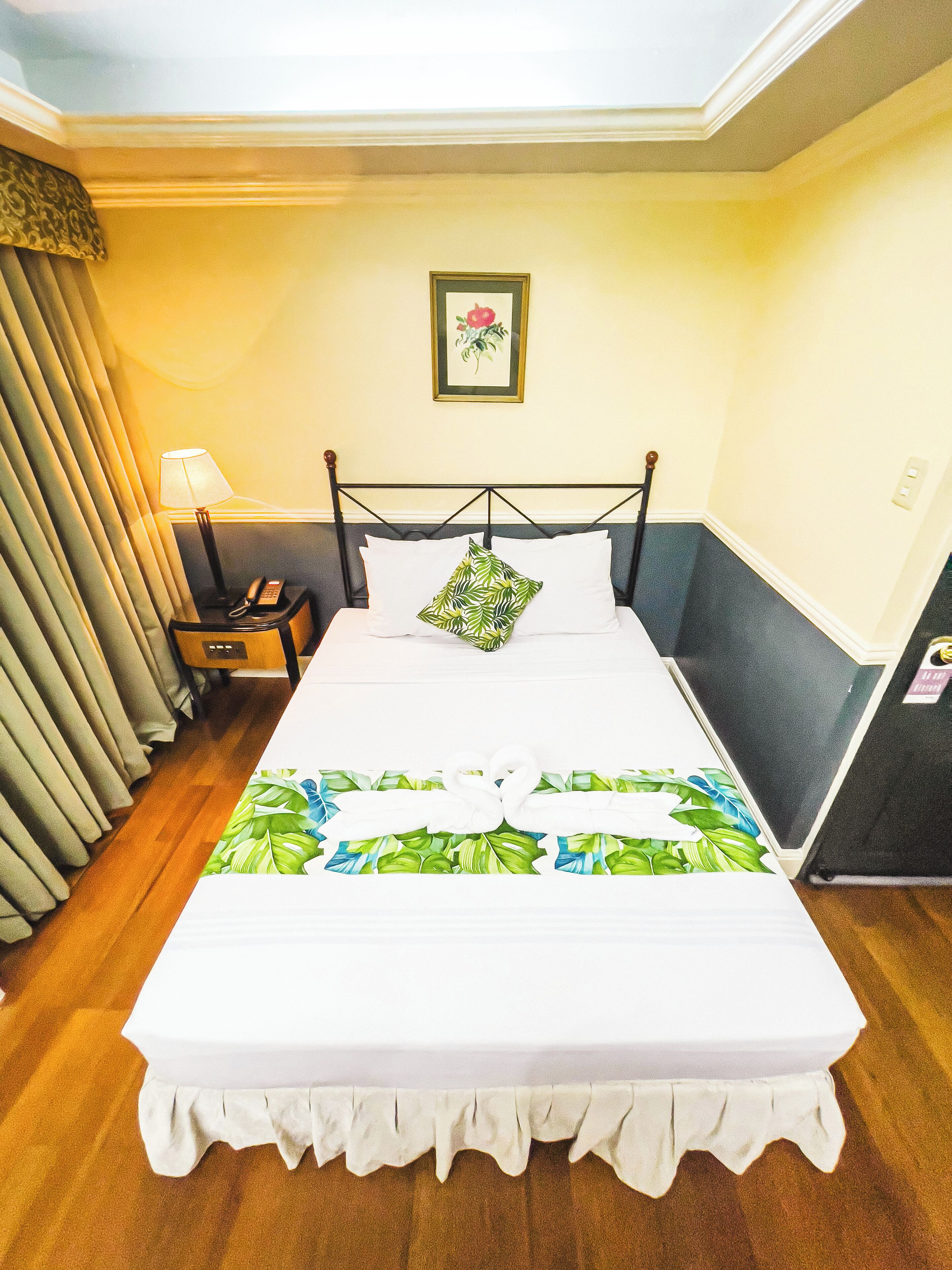 A neatly made bed with a white bedspread and a tropical leaf pattern, a bedside lamp, a framed picture on the wall, and a wooden nightstand.