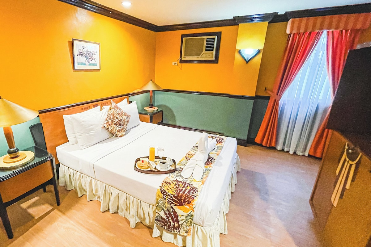 A hotel room with a bed, a bedside table, a chair, a tray with breakfast items, and a window with curtains.