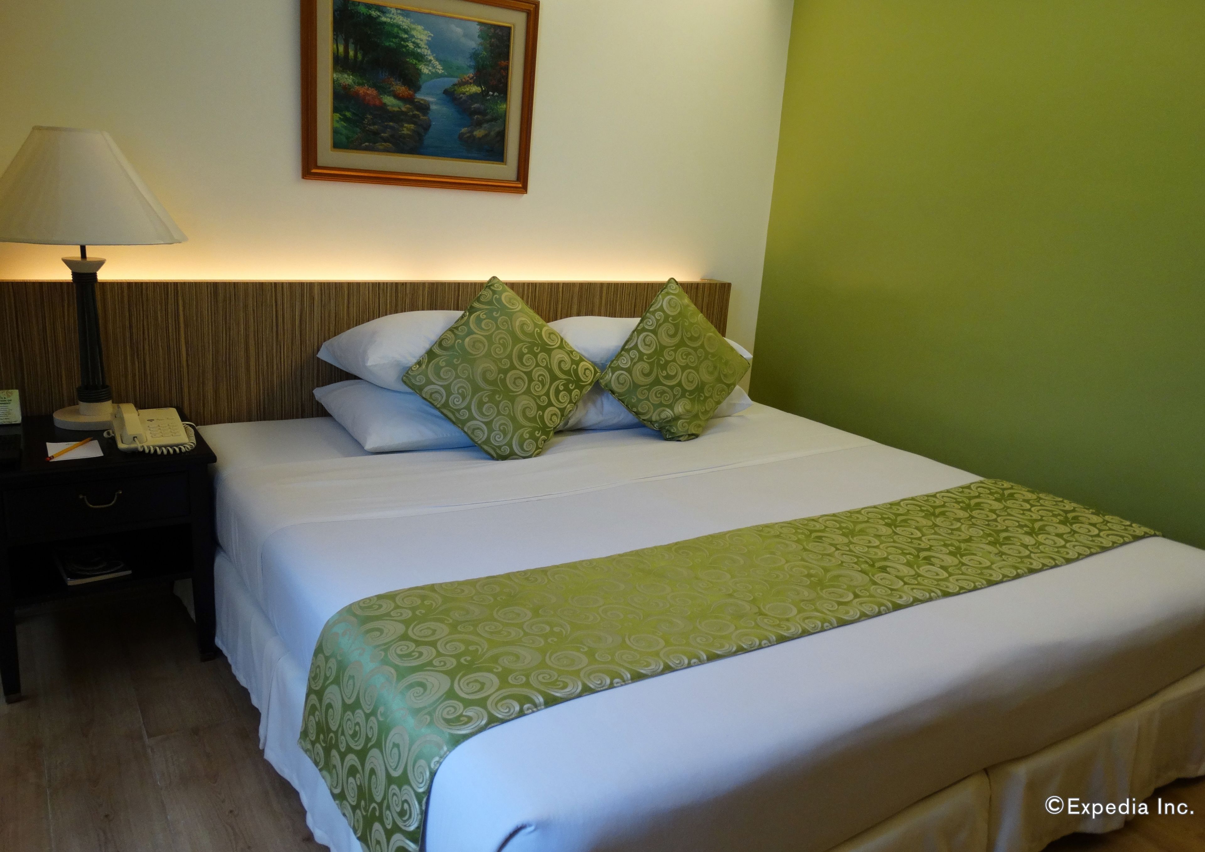 A neatly made bed with green patterned pillows and a green bedspread, a bedside lamp, a phone, and a framed painting on the wall.
