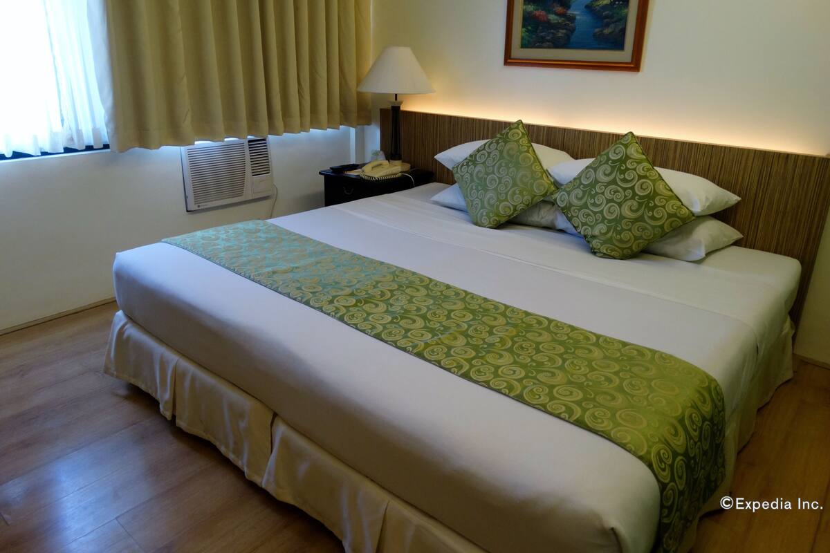 Una habitación de hotel con una cama grande, una mesita de noche con lámpara, un cuadro en la pared y una ventana con cortinas.