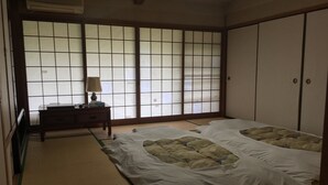 Ett traditionellt japanskt rum med tatamimattor på golvet, en futonmadrass, skjutdörrar och en liten byrå i trä med en lampa.