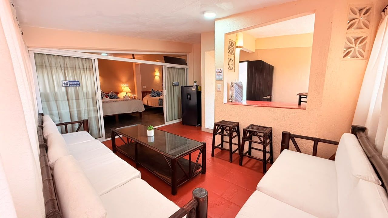 Un salon avec une table basse en verre, des canapés blancs, une petite table à manger avec des chaises et une porte coulissante donnant sur une chambre.
